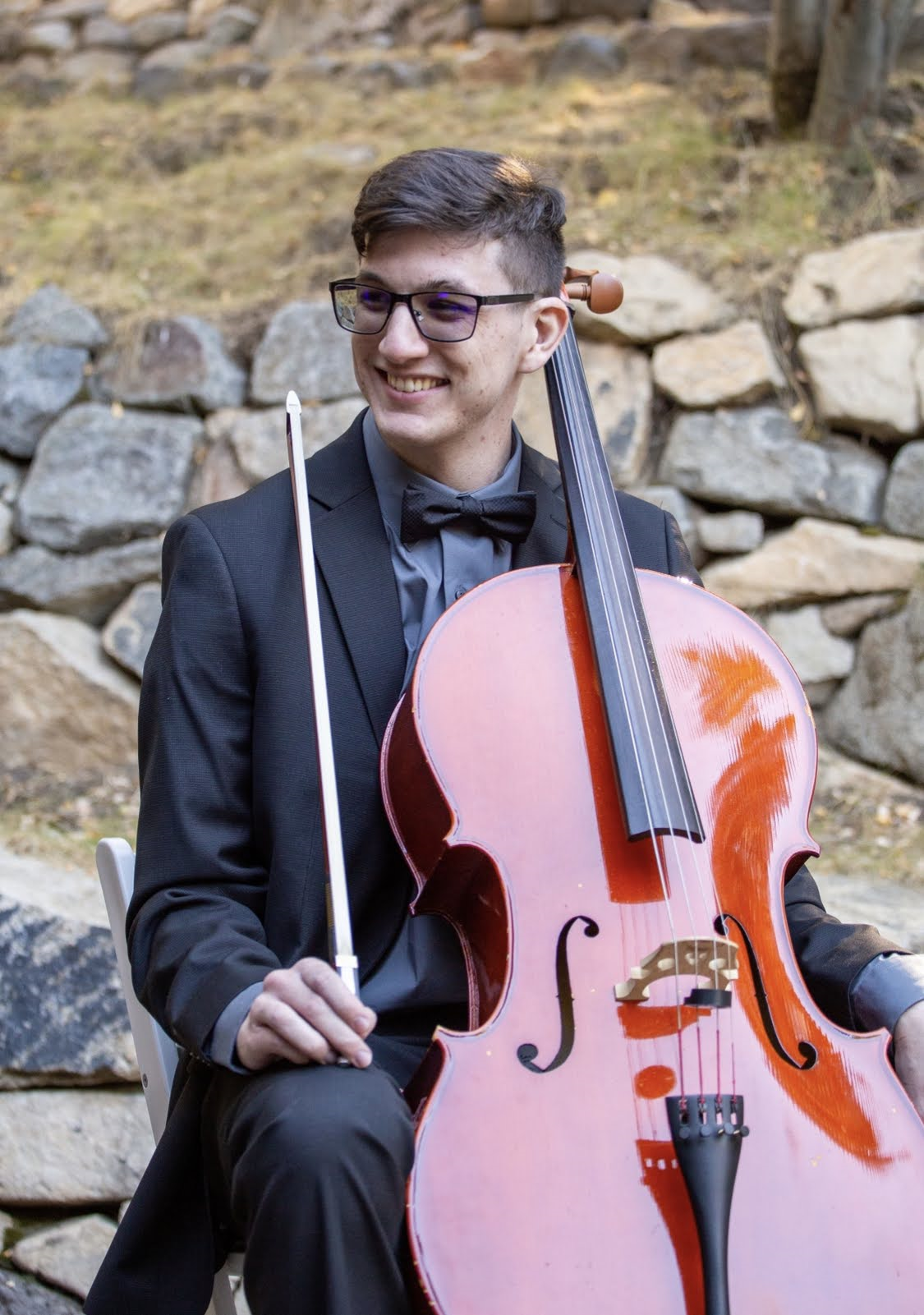 A picture of Cass Unterholzner sitting while playing the cello. They are holding their cello bow and have it resting on their knee upright. They are wearing a suit.